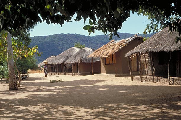 Row of traditional thatched mud huts in a rural Malawian village surrounded by hills and trees.