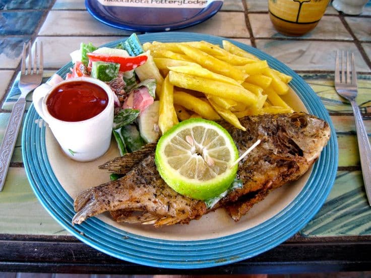 A plate of grilled chambo fish served with French fries, salad, and a slice of lime, alongside ketchup in a small cup.